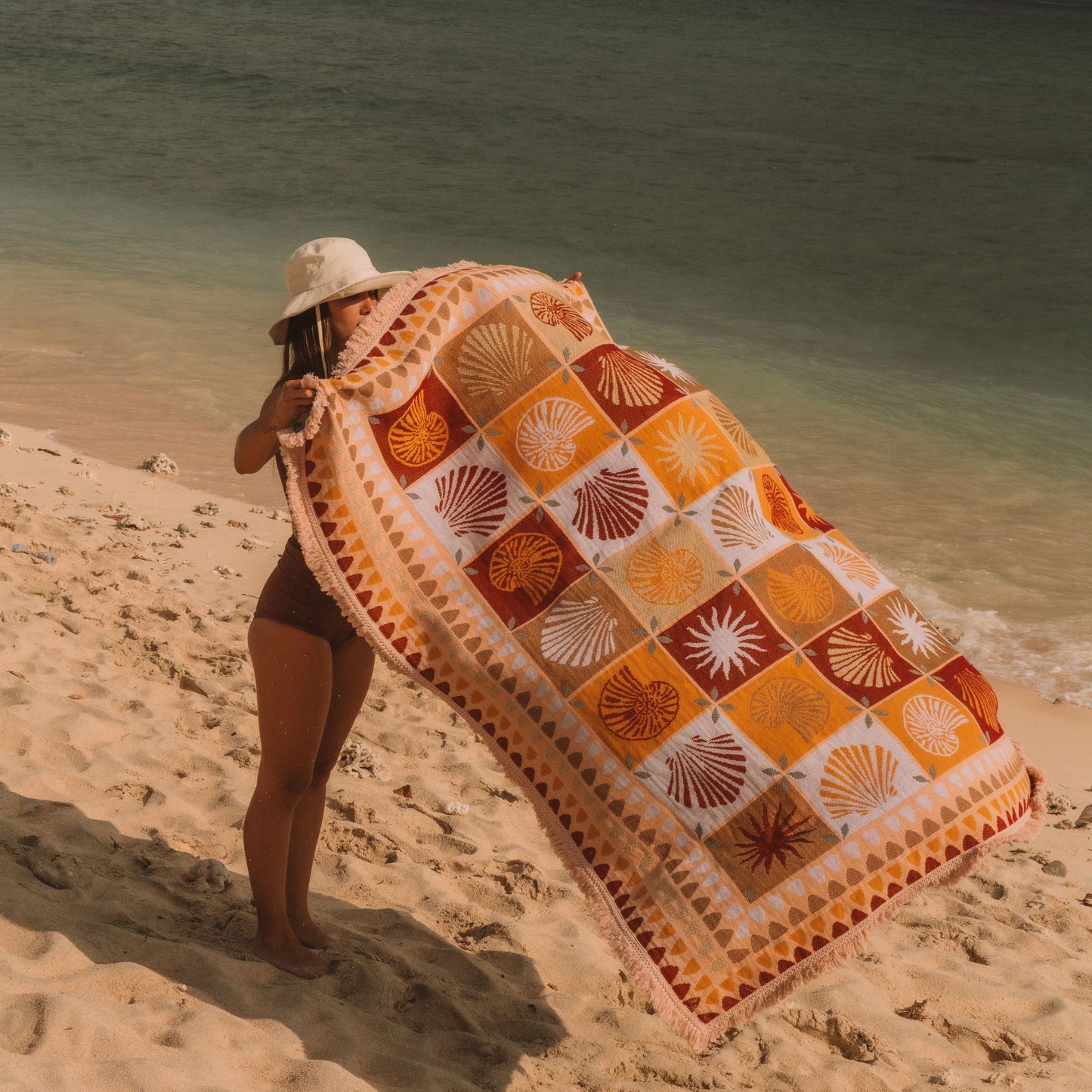 A woman on a sandy beach holds up a large patterned beach blanket or towel. The blanket features a retro patchwork design with orange, yellow, brown, and white sunburst and fan motifs.